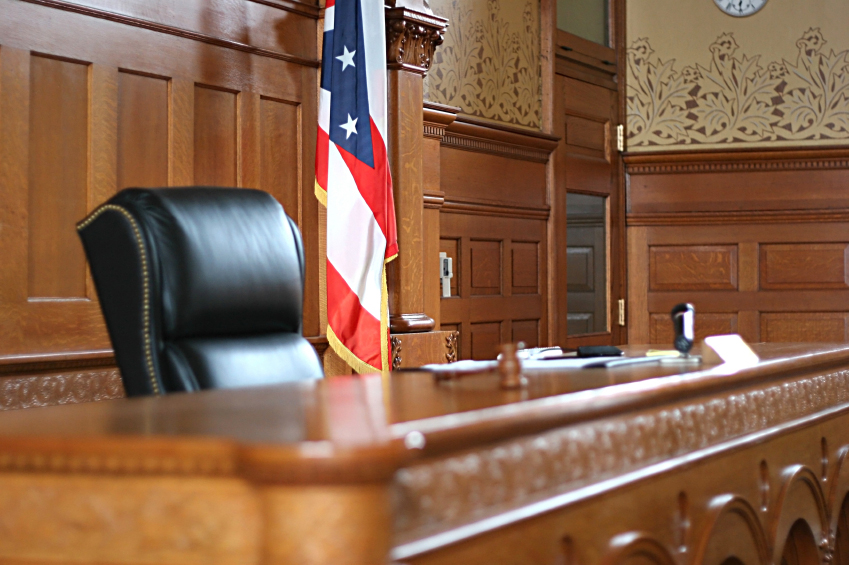 Empty judge's bench with a black leather chair and a flag in a wood-paneled courtroom.